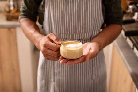 Cup of cappuccino. Close up of professional barista wearing striped apron holding cup of cappuccinoの写真素材