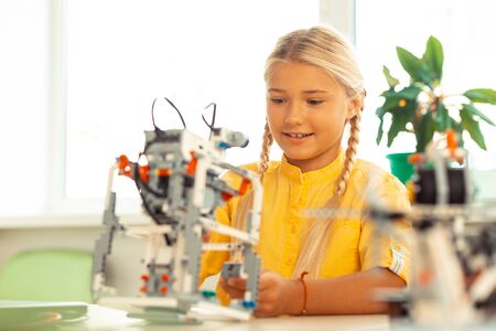 Modern school. Excited schoolgirl with long plaits sitting at her desk and building a robot of construction set at science lesson.の写真素材