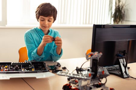 Young roboteer. Smiling schoolboy sitting at the school desk with computer on it and preparing for...