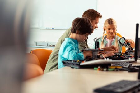 Interesting science. Smiling girl and concentrated boy building robots using construction set sitting with their teacher at their science lesson.の写真素材