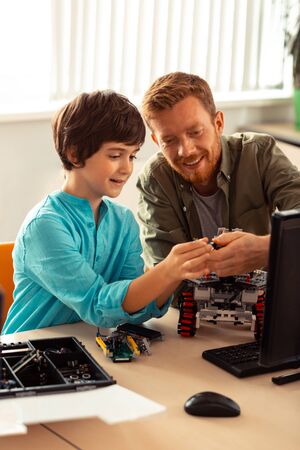 Showing the right way. Smiling science teacher helping his hard-working pupil with robot building sitting with him in computer lab.の写真素材
