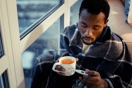 Modern technologies. Worried brunette male person holding cup with tea and looking at thermometer