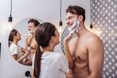 Smiling young woman putting shaving foam on cheerful face of her boyfriendの写真素材