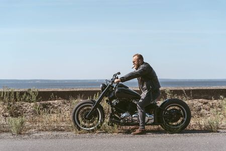 Sexy man in leather jacket posing on his bike by the oceanの写真素材