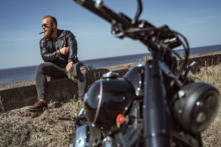 Attractive biker smoking sigar enjoying the view of the ocean by his bike