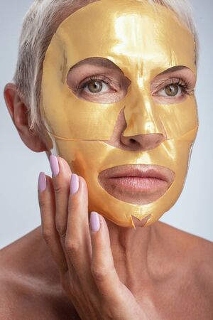 Gold mask. Delighted female pressing lips while putting mask on her faceの写真素材