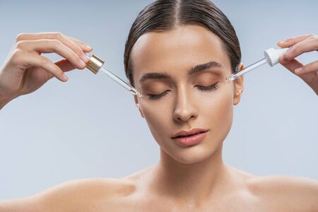 Double treatment. Beautiful woman holding two pipette near temples while taking care of her skin