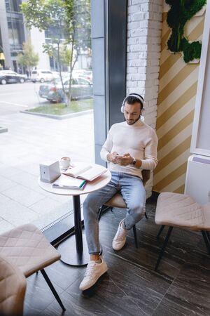 Kind bearded male person sitting near big window while looking at screen of his gadget