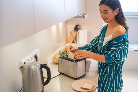 Cheerful young woman in sleepwear putting a slice of bread into the toaster