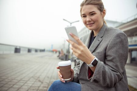 Smiling woman holding cup of coffee and reading message on cellphone stock photo. Website banner