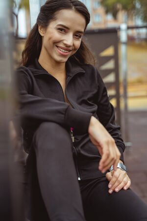 Pleased brunette female keeping smile on her face while sitting on the sports areaの写真素材