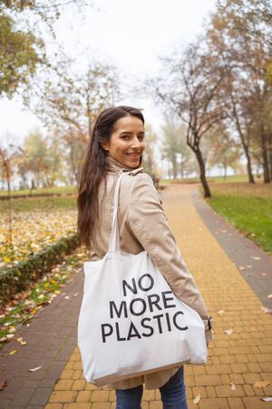 Eco bag. Pleased brunette keeping smile on her face while walking on the parkの写真素材