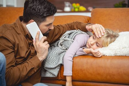 Worried man talking with doctor on cellphone while sick little girl lying on couch stock photoの写真素材