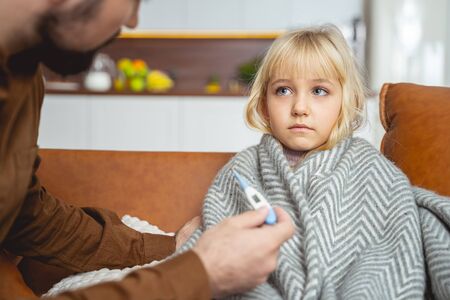 Cute little girl wrapped in blanket sitting on couch while her dad holding digital thermometer stock photoの写真素材