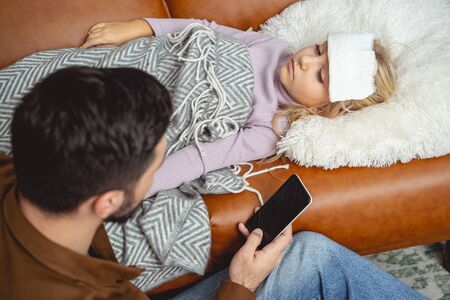 Young man holding smartphone while sitting beside his daughter with flu stock photo