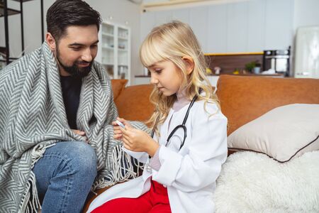 Adorable child holding digital thermometer while sitting with sick man on couch stock photoの写真素材