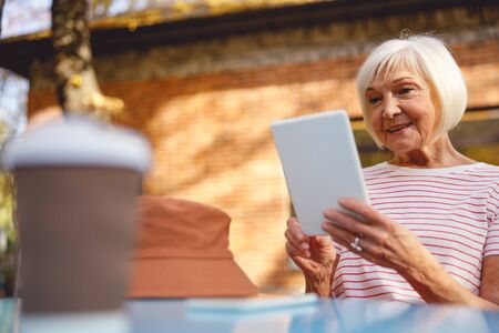 Cheerful olf lady sitting in the street cafe looking at her tablet