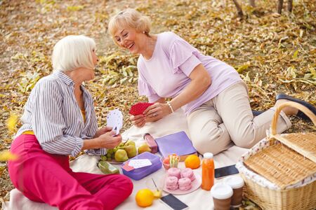 Two cheerful aged women playing cards and sitting on a picnicの写真素材