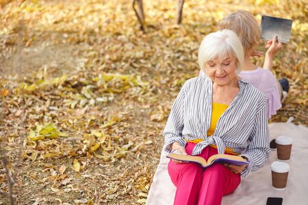 Two beautiful aged women sitting back to back on a blanket in the parkの写真素材