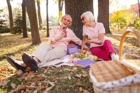 Two cheerful women laughing while sitting in the park on picnicの写真素材