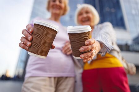 Close up picture of two coffee cups in hands of two cheerful old ladiesの写真素材