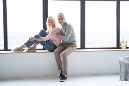 Positive man hugging his wife and sitting on a window seat near herの写真素材