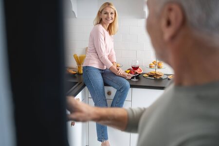 Beautiful lady smiling to husband while sitting on a kitchen countertop with food and wineの写真素材