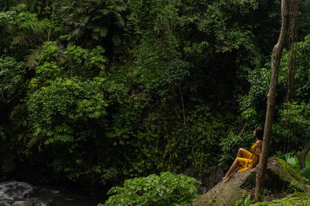 Pretty brunette girl sitting in semi position on big stone while looking at green plantsの写真素材
