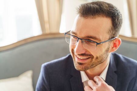 Closeup portrait of a pleased attractive Caucasian young male in spectacles touching his shirt...