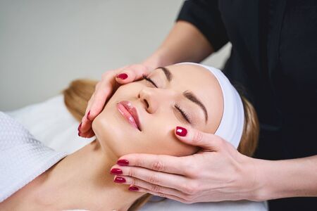 Happy girl is lying on couch with closed eyes while cosmetologist is taking care of her faceの写真素材