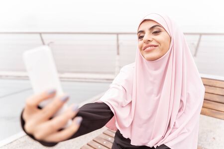 Cute Muslim lady sitting on a bench and smiling while talking photos of herselfの写真素材