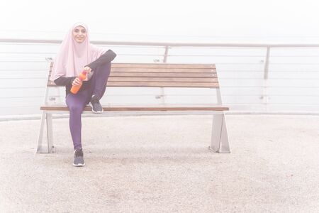 Smiling happy lady in hijab sitting comfortably outdoors with a sports bottle of water. Website...