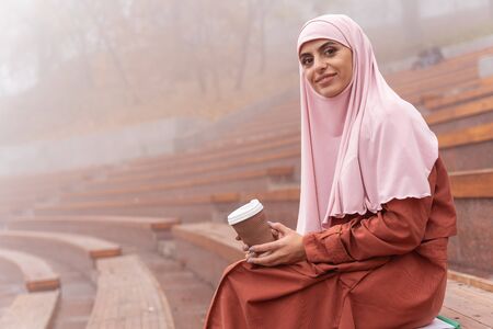 Beautiful Muslim lady with a paper cup of coffee sitting on the bench on grandstand