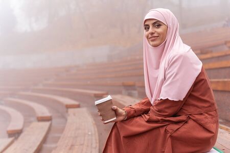 Pleased young woman in hijab relaxing on a grandstand with coffee to go. Website bannerの写真素材