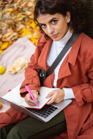 Beautiful young woman sitting near the tree with a laptop and making notes in her notebookの写真素材