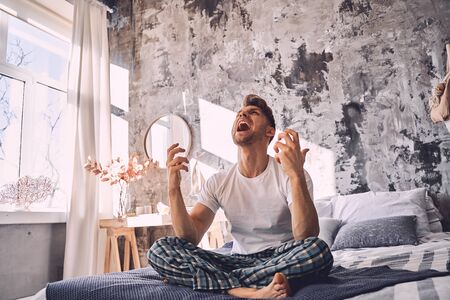 Emotional male person sitting on the bed and raising his arms while shouting in loudの写真素材