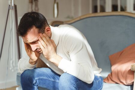Sad Caucasian male in a white pullover bending double with a pain on a sofa