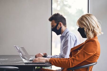 Side view of two concentrated Caucasian office workers in face masks working on their laptops