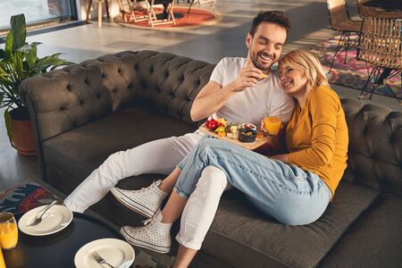 Breakfast time. Caring handsome man feeding his lovely young wife with a piece of cheeseの写真素材