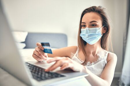 Young concentrated woman in mask buying some goods on Internet stock photo. Coronavirus conceptの写真素材