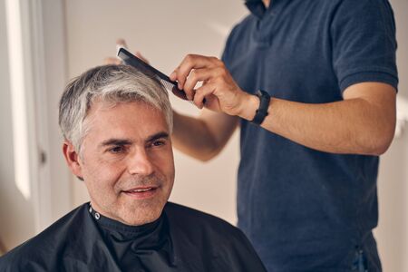 Elderly man sitting quietly while somebody wearing blue T-shirt and watch on his hand combs his hairの写真素材