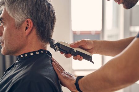Cropped photo of a Caucasian man sitting quietly while another male changing his hairstyle with help...