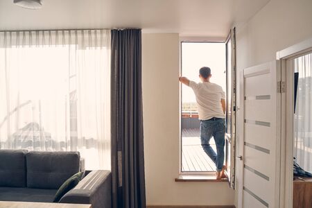 Male standing in his living room leaning on the wall and watching a nice view outsideの写真素材