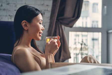 Close up of attractive brunette lady closing eyes and smiling while enjoying delicious hot drinkの写真素材