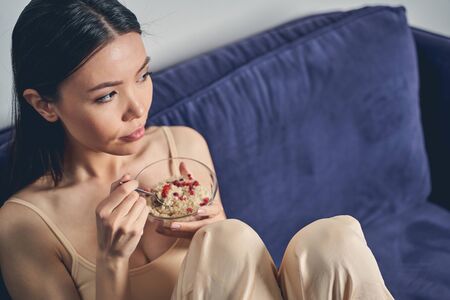 Beautiful brunette lady looking away with serous expression while holding glass bowl of delicious oat porridgeの写真素材