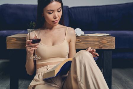 Attractive brunette lady sitting on the floor and holding glass of wine while studyingの写真素材