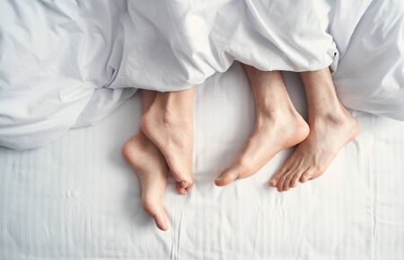 Top view of two pairs of bare feet lying on a white sheet in bed