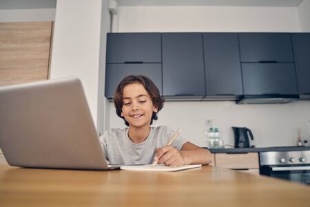 Smiling Caucasian boy spending time inside in the kitchen and using his computer while writing with pencilの写真素材