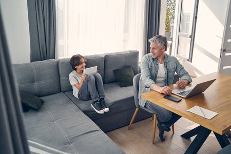 Grey haired adult man working at the table on laptop while his son playing online with tablet at homeの写真素材