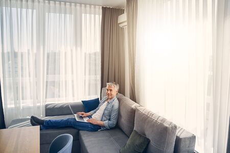 Smiling adult Caucasian man staying at home in cozy atmosphere while working on his laptopの写真素材